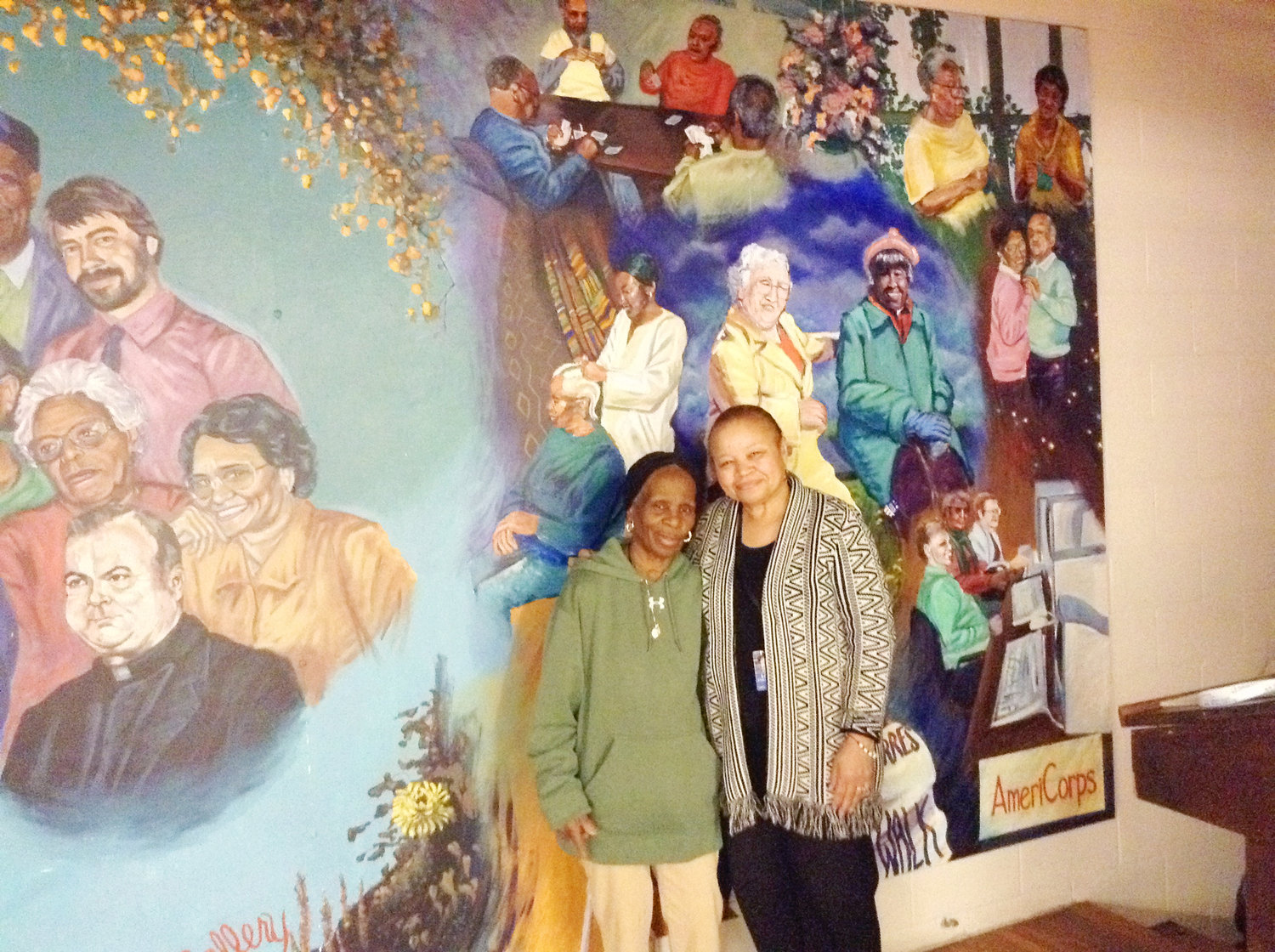 Lorraine DeCosta and Linda A’vant-Deishinni, director of the St. Martin de Porres Center, stand before a mural in the center’s dining hall. DeCosta, a stroke survivor, is one of many members of the St. Martin’s community to be depicted in the mural.